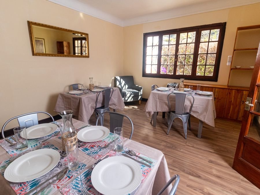 Comedor con mesas puestas con mantel, vajilla y flores, junto a un ventanal.